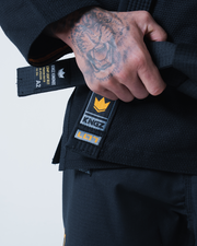 Kingz Legends Never Die Respect Gi in Black – close-up detail view: tattooed hand holding belt Kingz crown badge orange text black fabric texture