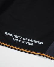 Kingz Legends Never Die Respect Youth Gi in Black – close-up detail view: white text "Respect Is Earned Not Given" orange stripe hem black fabric