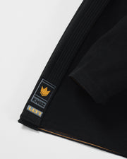 Kingz Legends Never Die Respect Women's Gi in Black – close-up detail view: black gi fabric yellow crown badge KINGZ logo LLTD text contrast