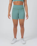 Kingz Kore Women's Training Shorts Green Light Sage - Full Body Front View Female Athlete Sage Mint Green Biker Style Short Cut High-Waist Wide Waistband Olive Green Contrast Stitching KINGZ White Text Right Outer Thigh Side Pocket Matching Sage Sports Bra White Sneakers Women's Training Shorts No-Gi BJJ Kore Collection [color:Light Green]